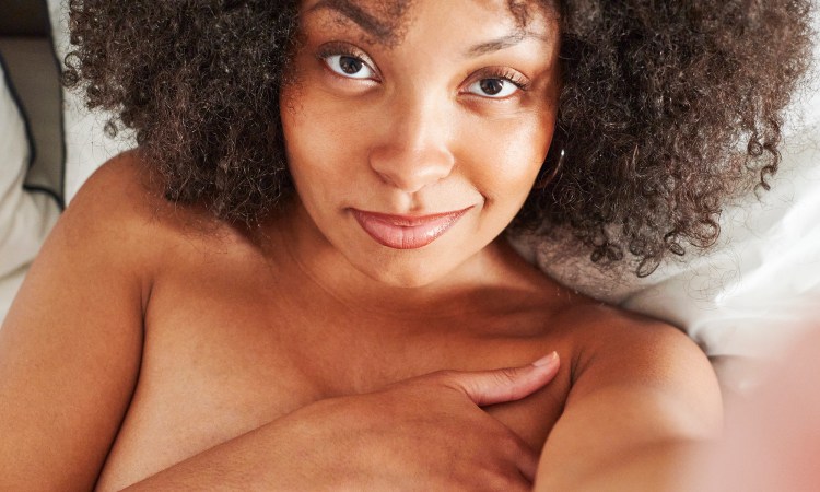 A Black woman takes a selfie where she smiles wryly at the camera. She is not wearing clothes and covers her chest with her hand. She has dark brown curly hair which frames her face, and is lying in a bed with white sheets.